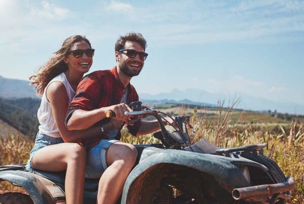 Off-Road Vehicle Insurance - Happy Young Couple Riding ATV Together Along a Dirt Road in the Countryside on a Sunny Day
